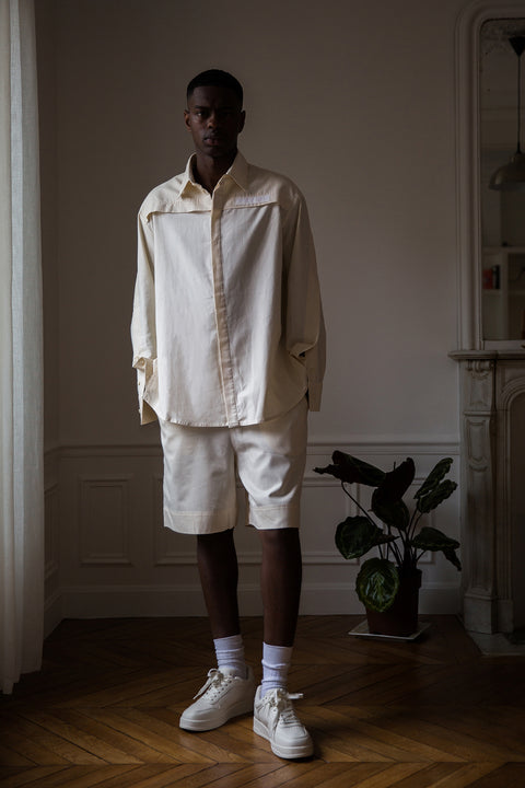 Thierno is wearing White Canvas sustainable natural beige color, organic cotton, genderless, inclusive, casual short, size M styled with a white tank top. The Essential short has minimal modern aesthetics design allowing comfort and bringing self-confidence. A limited-edition production is made locally in France to reduce environmental impact and guarantee high quality and durability of the products.
