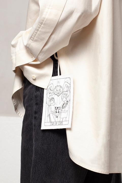 THE TWO OF CUPS tag is made from leftover deadstock white cotton poplin. The black and white tag has an exclusive design inspired by tarot cards drawn by the artist Maria Robla and is printed in collaboration with RoStudio. Wear the tag alone or attach it to the essential pieces to personalize them. The tag is sold with key holder in recycled plastic made in collaboration with Samji Atelier. White Canvas x Maria Robla.