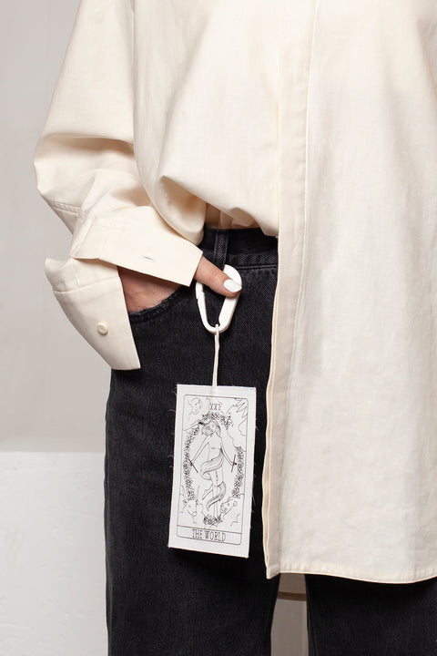 THE WORLD tag is made from leftover deadstock white cotton poplin. The black and white tag has an exclusive design inspired by tarot cards drawn by the artist Maria Robla and is printed in collaboration with RoStudio. Wear the tag alone or attach it to the essential pieces to personalize them. The tag is sold with key holder in recycled plastic made in collaboration with Samji Atelier. White Canvas x Maria Robla.