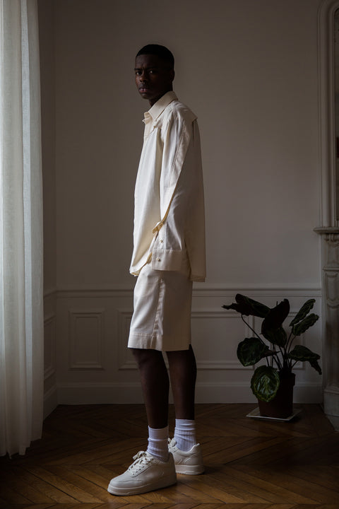 Thierno is wearing White Canvas sustainable natural beige color, organic cotton, genderless, inclusive, casual short, size M styled with a white tank top. The Essential short has minimal modern aesthetics design allowing comfort and bringing self-confidence. A limited-edition production is made locally in France to reduce environmental impact and guarantee high quality and durability of the products. 