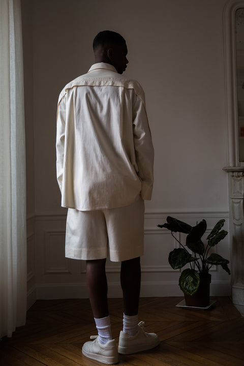 Thierno is wearing White Canvas sustainable natural beige color, organic cotton, genderless, inclusive, casual short, size M styled with a white tank top. The Essential short has minimal modern aesthetics design allowing comfort and bringing self-confidence. A limited-edition production is made locally in France to reduce environmental impact and guarantee high quality and durability of the products. 