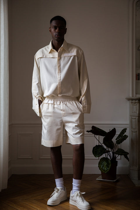 Thierno is wearing White Canvas sustainable natural beige color, organic cotton, genderless, inclusive, casual short, size M styled with a white tank top. The Essential short has minimal modern aesthetics design allowing comfort and bringing self-confidence. A limited-edition production is made locally in France to reduce environmental impact and guarantee high quality and durability of the products. 