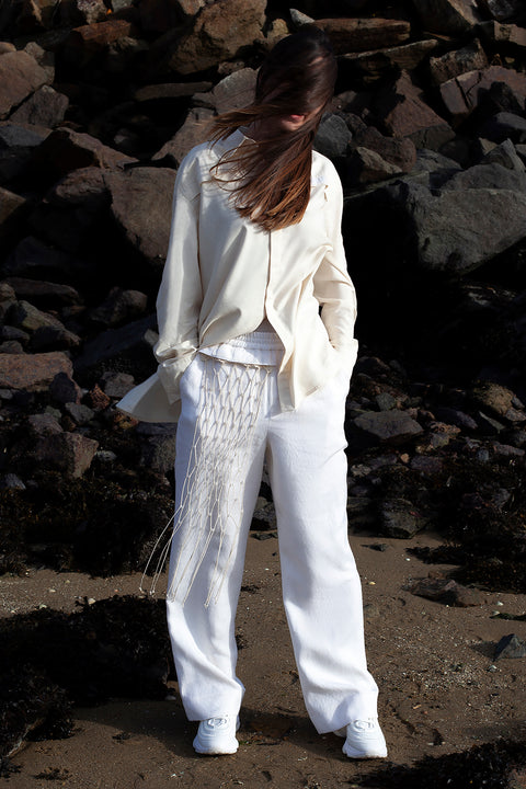 Romane is wearing the White Canvas ESSENTIAL SHIRT AND TROUSERS styled with the net vertical panel with recycled beads made from leftover deadstock fabrics. The net vertical panel with recycled beads is an exclusive unique sustainable piece made in France. It is made to be attached to the essential pieces to personalize them and wear them your unique way.