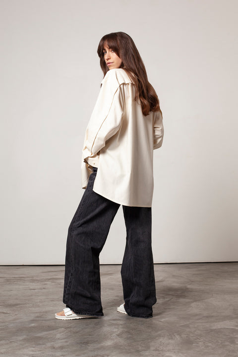 Léa is wearing White Canvas sustainable natural beige color, organic cotton oversized, genderless, inclusive, seasonless casual shirt, size S/M styled with black jeans. The Essential shirt has minimal modern aesthetics design allowing comfort and bringing self-confidence. A limited-edition production is made locally in France to reduce environmental impact and guarantee high quality and durability of the products. 