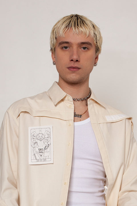 Clément is wearing the White Canvas ESSENTIAL SHIRT styled with THE TWO OF CUPS tag made from leftover deadstock white cotton poplin. The black and white tag has an exclusive design inspired by tarot cards drawn by the artist Maria Robla and is printed in collaboration with RoStudio. Wear the tag alone or attach it to the essential pieces to personalize them. The tag is sold with key holder in recycled plastic made in collaboration with Samji Atelier.  White Canvas x Maria Robla.