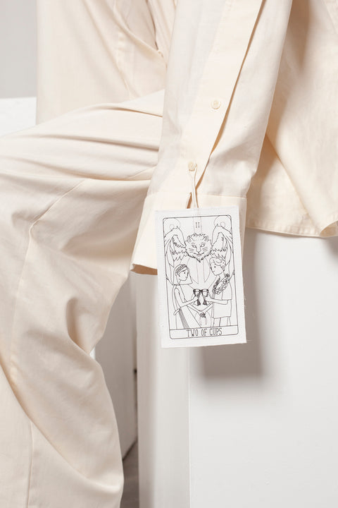 Léa is wearing the White Canvas ESSENTIAL SHRT styled with THE TWO OF CUPS tag made from leftover deadstock white cotton poplin. The black and white tag has an exclusive design inspired by tarot cards drawn by the artist Maria Robla and is printed in collaboration with RoStudio. Wear the tag alone or attach it to the essential pieces to personalize them. The tag is sold with key holder in recycled plastic made in collaboration with Samji Atelier. White Canvas x Maria Robla.
