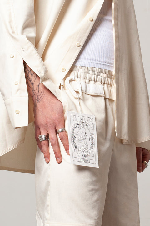 Clément is wearing the White Canvas ESSENTIAL SHORT styled with THE WORLD tag made from leftover deadstock white cotton poplin. The black and white tag has an exclusive design inspired by tarot cards drawn by the artist Maria Robla and is printed in collaboration with RoStudio. Wear the tag alone or attach it to the essential pieces to personalize them. The tag is sold with key holder in recycled plastic made in collaboration with Samji Atelier. White Canvas x Maria Robla.
